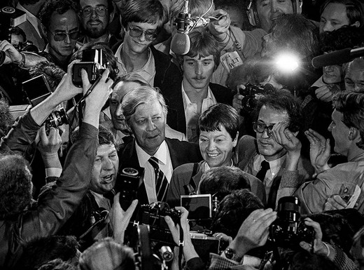 Die Abbildung zeigt eine Schwarz-weiß-Fotoaufnahme von Michael Jostmeier. Das Foto zeigt den Bundeskanzler Helmut Schmidt mit seiner Ehefrau gedrängt umringt von fotografierenden zahlreichen Journalisten.