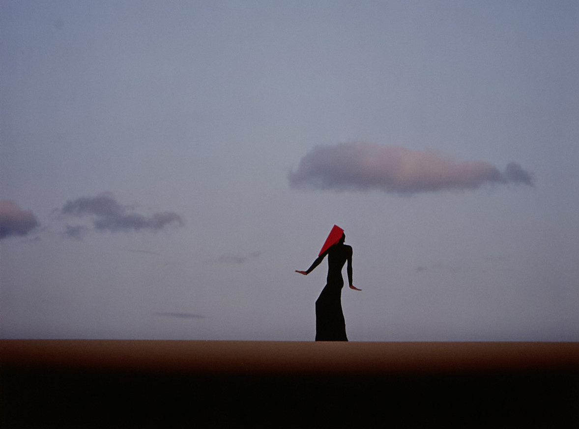 Eine einzelne Person mit langem Kleid und rotem Tuch über dem Kopf steht aufrecht in weiter Landschaft. Der Himmel ist klar und hell, mit wenigen, locker verteilten Wolken. Die Szene wirkt minimalistisch, der Horizont verläuft gerade und teilt das Bild in zwei Flächen.