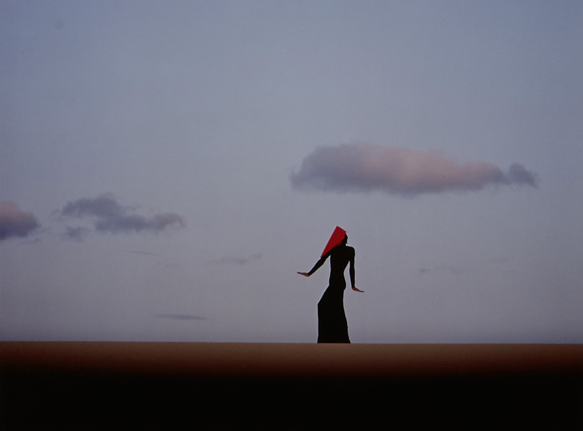 Eine einzelne Person mit langem Kleid und rotem Tuch über dem Kopf steht aufrecht in weiter Landschaft. Der Himmel ist klar und hell, mit wenigen, locker verteilten Wolken. Die Szene wirkt minimalistisch, der Horizont verläuft gerade und teilt das Bild in zwei Flächen.