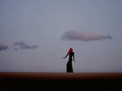 Eine einzelne Person mit langem Kleid und rotem Tuch über dem Kopf steht aufrecht in weiter Landschaft. Der Himmel ist klar und hell, mit wenigen, locker verteilten Wolken. Die Szene wirkt minimalistisch, der Horizont verläuft gerade und teilt das Bild in zwei Flächen.