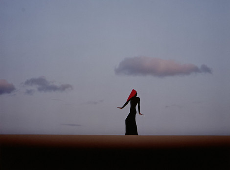 Eine einzelne Person mit langem Kleid und rotem Tuch über dem Kopf steht aufrecht in weiter Landschaft. Der Himmel ist klar und hell, mit wenigen, locker verteilten Wolken. Die Szene wirkt minimalistisch, der Horizont verläuft gerade und teilt das Bild in zwei Flächen.