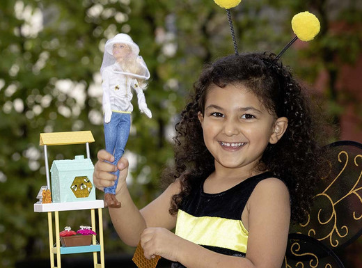 Die Abbildung zeigt ein junges Mädchen in einem Bienenkostüm.  Sie hält eine Barbiepuppe mit Imkeroutfit in der Hand und lächelt in die Kamera. Die Fotoaufnahme stammt von Marie Theres Graf.