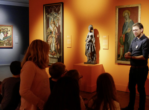 Die Abbildung zeigt eine Gruppe von Schüler*innen beim Besuch einer Ausstellung im Germanischen Nationalmuseum. Ein Mann steht vor einem Gemälde und erläutert es. Die Schüler*innen sitzen im Halbkreis um ihn und das Kunstwerk herum und hören zu. Die Fotoaufnahme stammt von Thomas Ruppenstein.