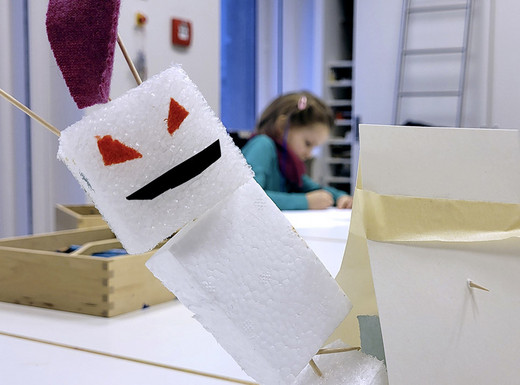 Im Vordergrund steht eine aus weißen Schaumstoffstücken, Zahnstochern und Papier gebastelte Figur mit roten, dreieckigen Augen und einem schrägen Mund. Die Figur trägt ein lilafarbenes Element am Kopf. Im Hintergrund sind ein Tisch, eine Holzbox, eine Leiter und weitere Bastelmaterialien zu sehen. Die Szene spielt in einem hellen Raum mit weißen Wänden.