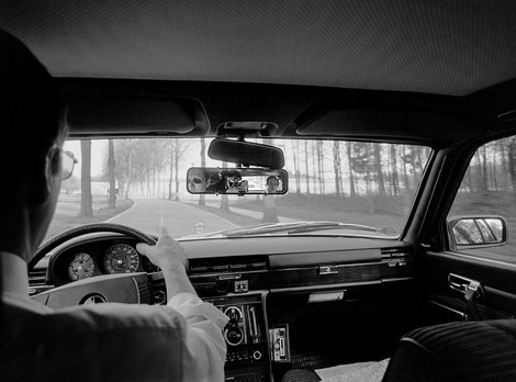 Die Abbildung zeigt eine Schwarz-weiß-Fotoaufnahme von Michael Jostmeier. Das Foto ist von der Rücksitzbank eines Autos während der Fahrt aufgenommen. Im Rückspiegel kann man den Fotograf und den Ministerpräsidenten Johannes Rau erkennen.