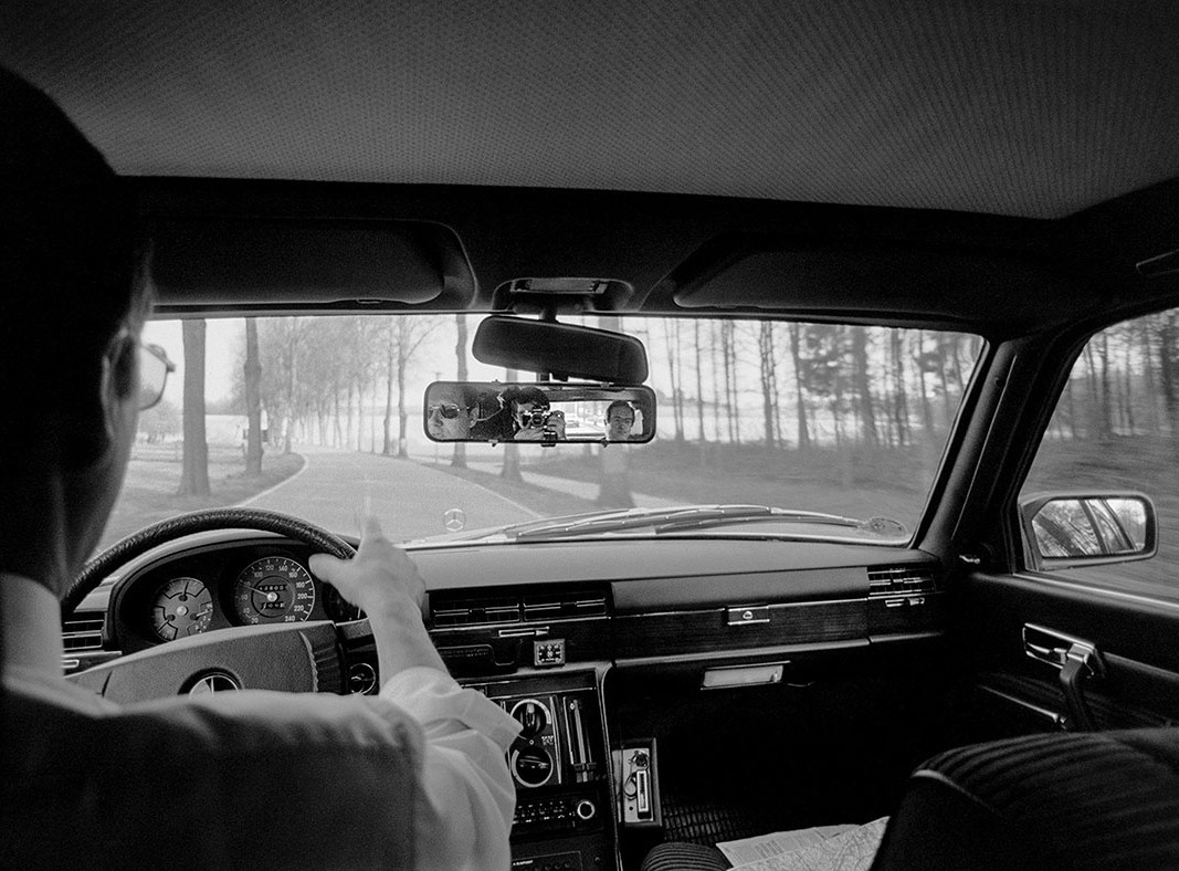 Die Abbildung zeigt eine Schwarz-weiß-Fotoaufnahme von Michael Jostmeier. Das Foto ist von der Rücksitzbank eines Autos während der Fahrt aufgenommen. Im Rückspiegel kann man den Fotograf und den Ministerpräsidenten Johannes Rau erkennen.