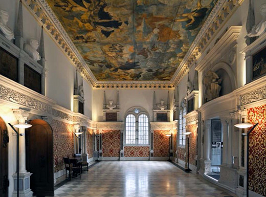 Die Fotoaufnahme zeigt den prunkvollen Hirsvogelsaal mit hoher, reich verzierter Decke, die ein großflächiges Deckenfresko zeigt. Die Wände sind mit roten, goldgemusterten Wandteppichen und weißen Stuckelementen geschmückt. Große Fenster lassen Tageslicht herein, das den Marmorboden und die kunstvollen Details beleuchtet. An den Seiten stehen Skulpturen und Büsten auf Konsolen, während Wandlampen warmes Licht spenden. Die Fotoaufnahme stammt von Uwe Niklas.