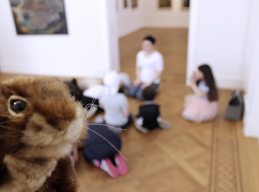 Kindergartengruppe und Elli Eichhörnchen in der Kunstvilla | Fotoaufnahme von Thomas Ruppenstein Die Abbildung zeigt im Hintergrund eine Kindergartengruppe in der Kunstvilla. Die Plüsch-Handpuppe Elli Eichhörnchen schaut im Vordergrund von links in die Kamera hinein. Die Fotoaufnahme stammt von Thomas Ruppenstein.