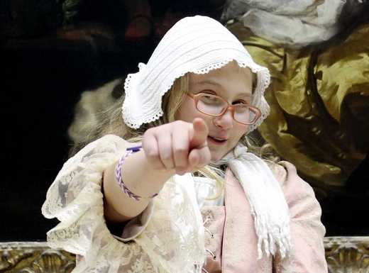 Die Fotoaufnahme zeigt ein Mädchen in historischer Verkleidung vor einem Gemälde des Germanischen Nationalmuseums. Es zeigt mit seiner rechten Hand freudig auf die Betrachtenden. Die Fotoaufnahme stammt von Thomas Ruppenstein.