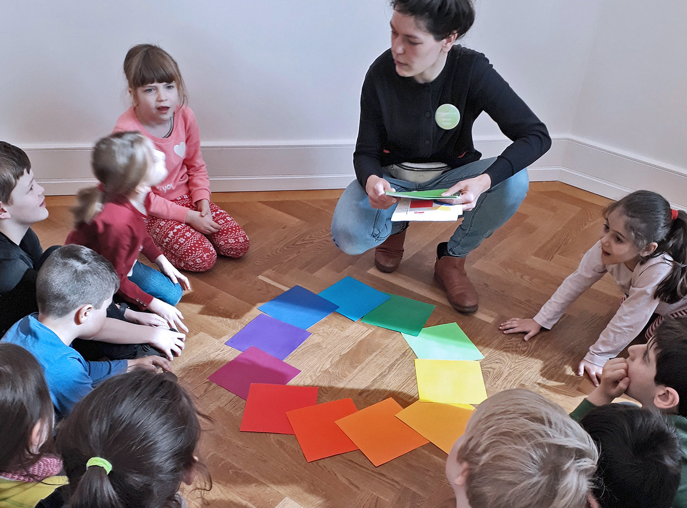 Die Abbildung zeigt Kinder einer Grundschulklasse, die sich im Kreis sitzend über am Boden liegende Farbbeispiele austauschen. Die Fotoaufnahme stammt von M. F.