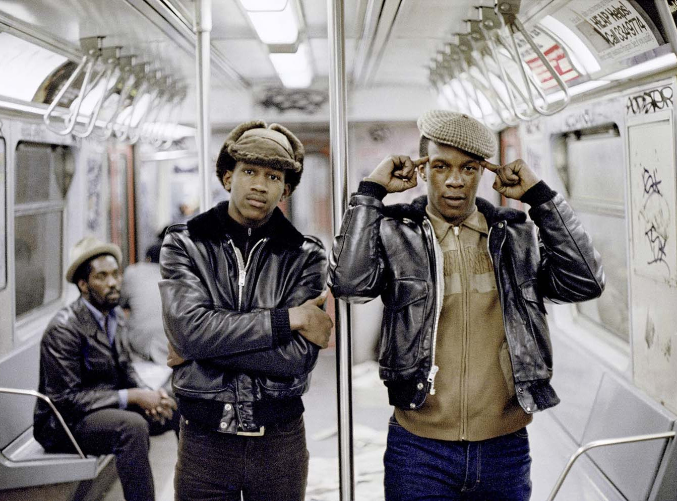 Die Abbildung zeigt eine Fotografie von Jamel Shabazz aus dem Jahr 1981. Zwei junge afroamerikanische Männer mit dunklen Lederjacken und Schirmmützen stehen in einem New Yorker U-Bahn-Wagen. Einer verschränkt die Arme, der andere hält sich die Zeigefinger an den Kopf. Im Hintergrund sitzt ein weiterer afroamerikanischer Mann auf einer Bank. Die Wände und Türen des Wagens sind mit Graffiti bedeckt.