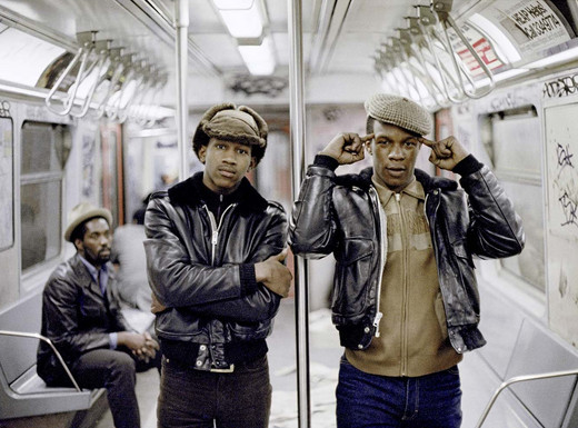 Die Abbildung zeigt eine Fotografie von Jamel Shabazz aus dem Jahr 1981. Zwei junge afroamerikanische Männer mit dunklen Lederjacken und Schirmmützen stehen in einem New Yorker U-Bahn-Wagen. Einer verschränkt die Arme, der andere hält sich die Zeigefinger an den Kopf. Im Hintergrund sitzt ein weiterer afroamerikanischer Mann auf einer Bank. Die Wände und Türen des Wagens sind mit Graffiti bedeckt.