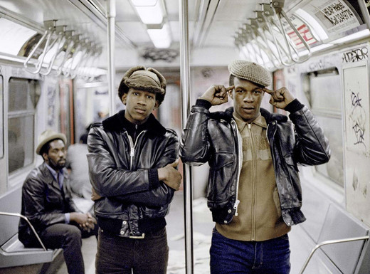 Die Abbildung zeigt eine Fotografie von Jamel Shabazz aus dem Jahr 1981. Zwei junge afroamerikanische Männer mit dunklen Lederjacken und Schirmmützen stehen in einem New Yorker U-Bahn-Wagen. Einer verschränkt die Arme, der andere hält sich die Zeigefinger an den Kopf. Im Hintergrund sitzt ein weiterer afroamerikanischer Mann auf einer Bank. Die Wände und Türen des Wagens sind mit Graffiti bedeckt.