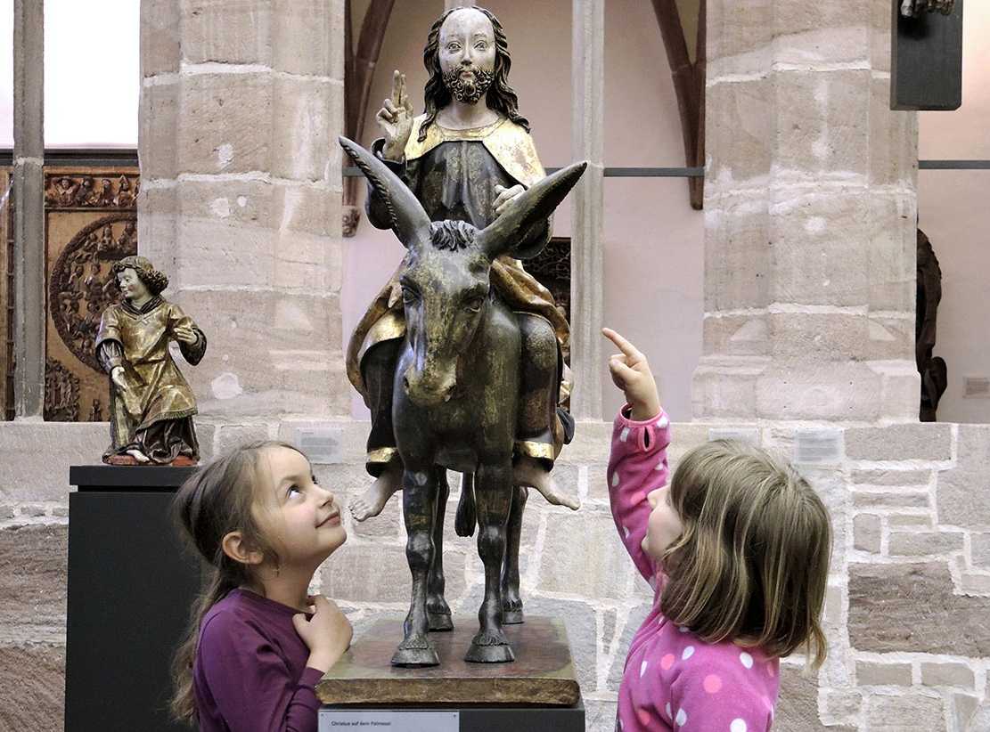 Die Abbildung zeigt zwei Kinder vor der Figur „Christus auf dem Palmesel“, die etwa zwischen 1370 und 1380 entstanden ist. Die Fotoaufnahme stamm von Thomas Ruppenstein.