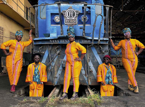 Direkt aus Lomé: Die Musikgruppe Nana Benz du Togo fasziniert mit ihrem einzigartig-unkonventionellen Sound. Fotoaufnahme von Arnold Anani. Die Abbildung zeigt fünf Personen der Musikgruppe "Nana Benz du Togo" in grell-orangen Oberalls vor einer alten blauen Diesellokomotive. Die Fotoaufnahme stammt von Arnold Anani.