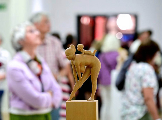 Besucher*innen bei der Eröffnung der Ausstellung zum NN-Kunstpreis 2025 | Fotoaufnahme von Joachim Winkler (NN) Die Abbildung zeigt im Vordergrund eine kleine Holzskulptur. Es handelt sich um eine sich bückende nackte Frau, auf deren Rücken ein Baby krabbelt. Im Hintergrund sind Personen bei der Betrachtung von anderen Kunstwerken zu sehen. Die Fotoaufnahme stammt von Joachim Winkler.