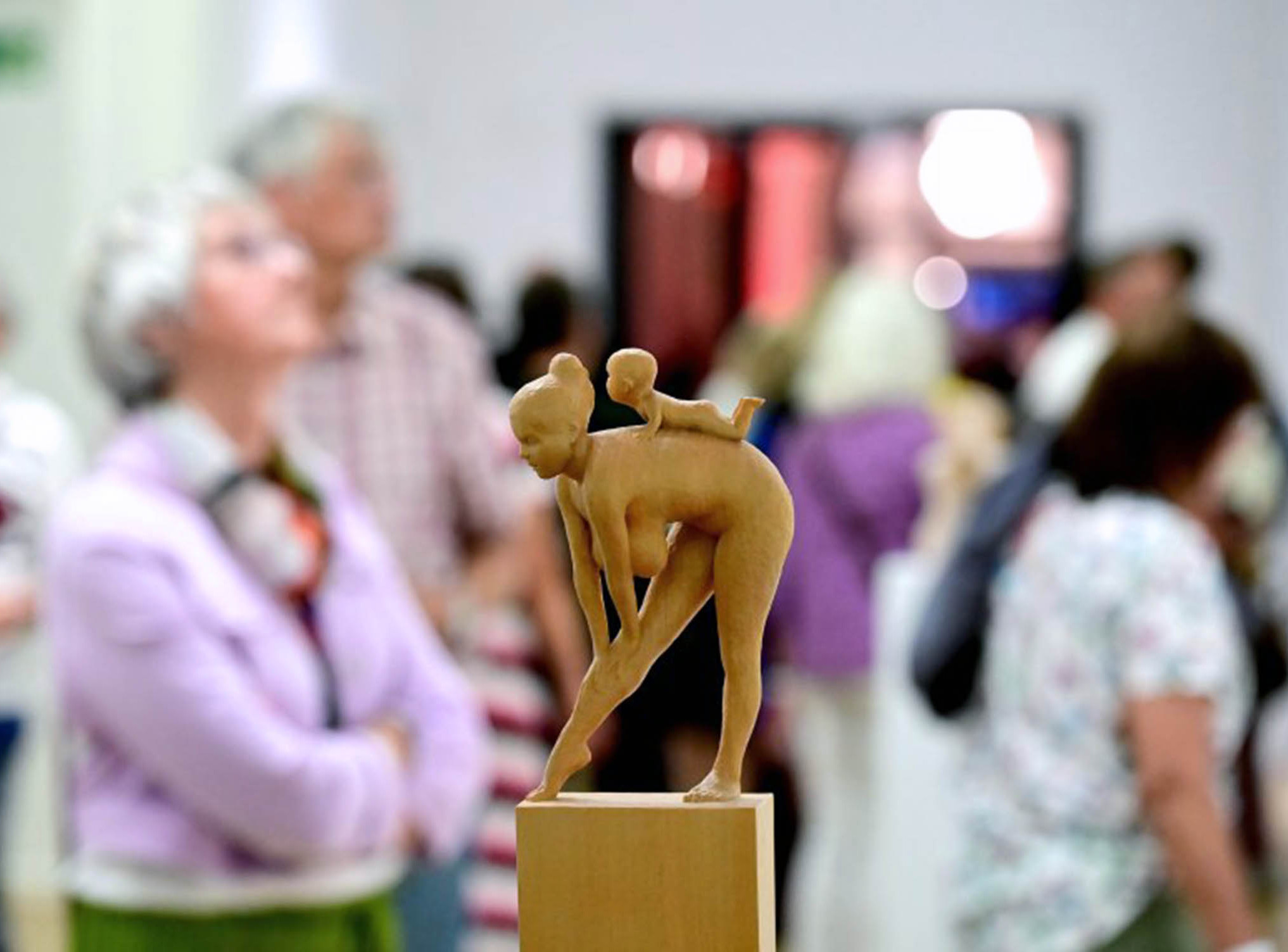 Besucher*innen bei der Eröffnung der Ausstellung zum NN-Kunstpreis 2025 | Fotoaufnahme von Joachim Winkler (NN) Die Abbildung zeigt im Vordergrund eine kleine Holzskulptur. Es handelt sich um eine sich bückende nackte Frau, auf deren Rücken ein Baby krabbelt. Im Hintergrund sind Personen bei der Betrachtung von anderen Kunstwerken zu sehen. Die Fotoaufnahme stammt von Joachim Winkler.