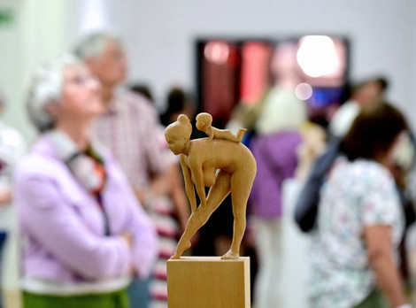 Die Abbildung zeigt im Vordergrund eine kleine Holzskulptur. Es handelt sich um eine sich bückende nackte Frau, auf deren Rücken ein Baby krabbelt. Im Hintergrund sind Personen bei der Betrachtung von anderen Kunstwerken zu sehen. Die Fotoaufnahme stammt von Joachim Winkler.