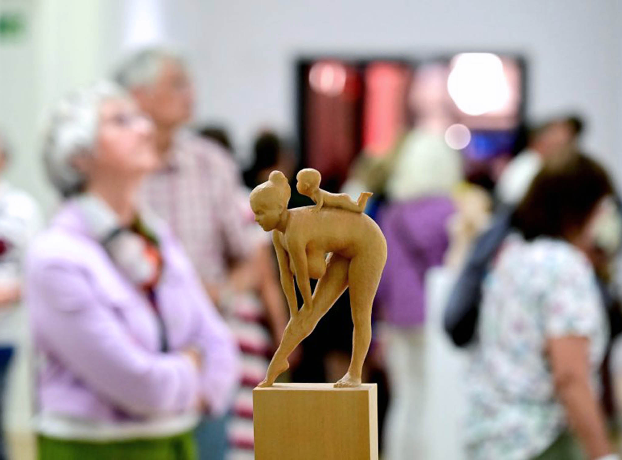 Besucher*innen bei der Eröffnung der Ausstellung zum NN-Kunstpreis 2025 | Fotoaufnahme von Joachim Winkler (NN) Die Abbildung zeigt im Vordergrund eine kleine Holzskulptur. Es handelt sich um eine sich bückende nackte Frau, auf deren Rücken ein Baby krabbelt. Im Hintergrund sind Personen bei der Betrachtung von anderen Kunstwerken zu sehen. Die Fotoaufnahme stammt von Joachim Winkler.