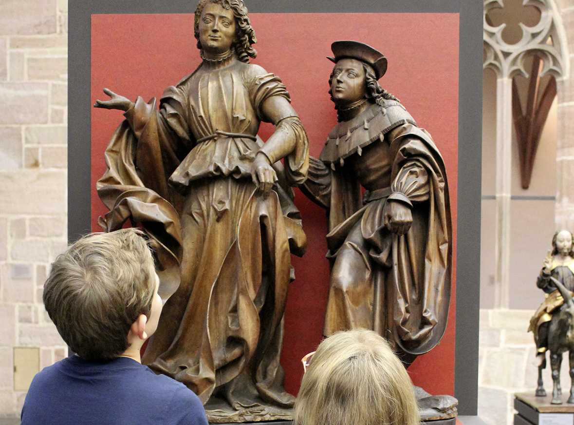 Die Abbildung zeigt zwei Kinder vor der Figurengruppe „Erzengel Raphael und der junge Tobias“. Die aus Holz geschnitzte Figurengruppe wurde von Veit Stoß im Jahr 1516 gefertigt. Die Fotoaufnahme stammt von Thomas Ruppenstein.